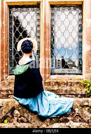 Una donna con un vestito blu e un cappello di paglia è seduta su un muro di pietra, guardando fuori dalla finestra. Foto Stock