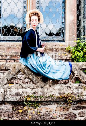 Una donna in abiti d'epoca siede su un muro di pietra di fronte a una finestra. Foto Stock