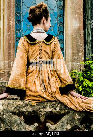 Una donna in costume storico siede su una sporgenza di pietra, la schiena alla telecamera. Indossa un lungo abito color oro con finiture nere. Foto Stock