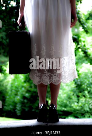 Una persona con un abito bianco e un tacco nero tiene una valigia, in piedi davanti a uno sfondo verde. Foto Stock