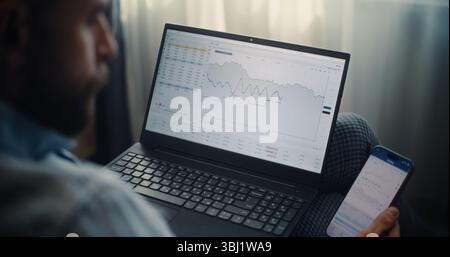 Primo piano di un esperto finanziario professionale che lavora su smartphone e laptop, monitoraggio delle azioni in tempo reale, grafici del mercato di Exchange nell'app di trading online. Uomo d'affari di successo nel comfort Home Office. Foto Stock