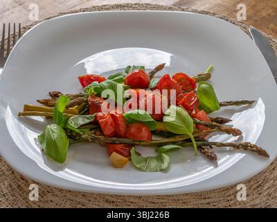Asparagi arrostiti conditi con succosi pomodori ciliegini e foglie di basilico, serviti su un piatto bianco pulito, freschi e sani. Foto Stock