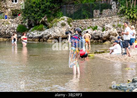 Taormina, Sizilien - Italia - 05-04-2025: Donna con fotocamera in piedi in acque poco profonde che scatta foto, circondata da turisti, costa rocciosa e vegetazione Foto Stock