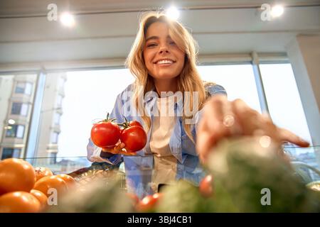 Donna felice che sceglie i pomodori maturi al supermercato. Vista ravvicinata di verdure fresche e clienti allegri in un abbigliamento casual. Concetto di mangiare sano Foto Stock