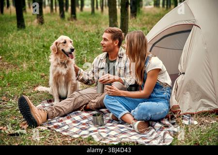 Una coppia gioiosa si imbarca in una deliziosa gita in campeggio insieme al loro amato Golden retriever, godendosi la serenità di una foresta tranquilla e creatina Foto Stock