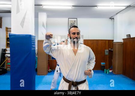 Atleta di karate che pratica tecniche di combattimento in un dojo, indossando un kimono bianco e una cintura marrone Foto Stock