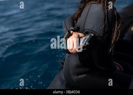 Il subacqueo indossa una muta si siede su una barca, tenendo in mano un computer per immersioni e preparandosi per un'avventura subacquea in mare Foto Stock