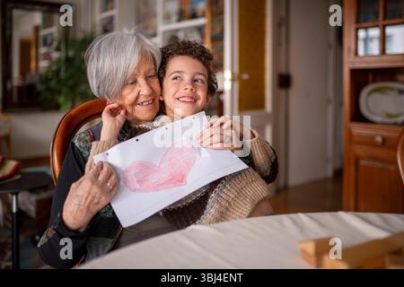 Nonna e nipote che si abbracciano e sorridono, mostrano un disegno di un cuore, seduti a un tavolo in salotto, godendo del tempo in famiglia insieme Foto Stock
