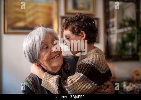 Nipote felice che sussurra segreti per sorridere la nonna, abbracciare e giocare insieme a casa, godendosi il tempo in famiglia Foto Stock