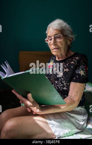 Una donna anziana è impegnata a leggere una cartella verde mentre è seduta comodamente su un letto. Foto Stock