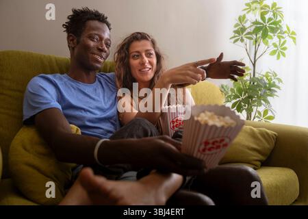 Una giovane coppia felice e variegata si rilassa su un divano a casa, mangiando popcorn e guardando un film insieme. Un'autentica scena di una serata cinematografica accogliente, lo Foto Stock