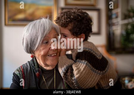 Nipote felice che sussurra segreti per sorridere nonna, godendo teneri momenti di affetto e complicità a casa Foto Stock
