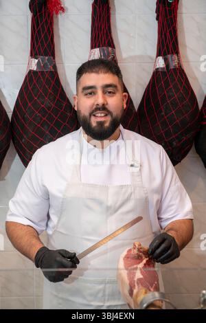 Macellaio con guanti neri che si prepara a affettare il prosciutto serrano con un coltello da prosciutto in una macelleria con prosciutti appesi Foto Stock