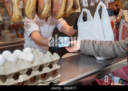 Macellaio che indossa guanti neri che ricevono banconote in euro dal cliente che acquista generi alimentari in macelleria con le gambe di prosciutto stagionato appese Foto Stock