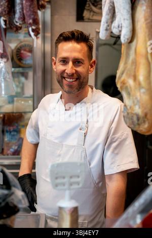 Ritratto di un macellaio sorridente nella sua macelleria, con prosciutto iberico e salsicce appese sullo sfondo Foto Stock