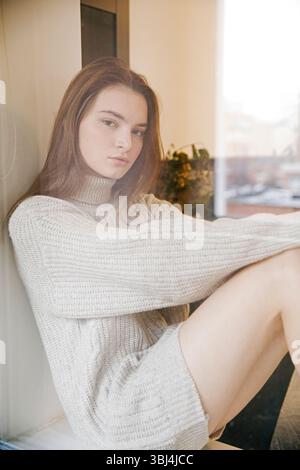 Giovane donna in maglione seduta sul davanzale Foto Stock