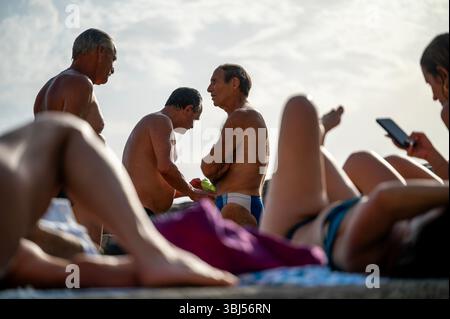 Tre uomini italiani più anziani si trovano sulla spiaggia di Vernazza, in Italia, incorniciati umoristicamente tra le gambe delle donne che prendono il sole in primo piano. Foto Stock