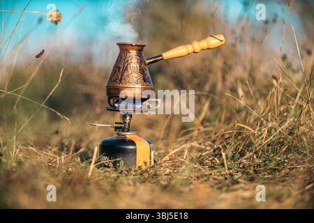 Il caffè viene preparato in una caffettiera incisa in rame sul bruciatore a gas contro il cielo sfocato blu. Concetto di relax ed escursioni in campagna in bicicletta Foto Stock