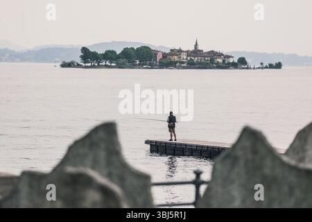 Pescatore su un molo di fronte all'Isola dei pescatori sul Lago maggiore, con edifici storici e campanile della chiesa visibili in lontananza, Piemonte, Italia. Foto Stock