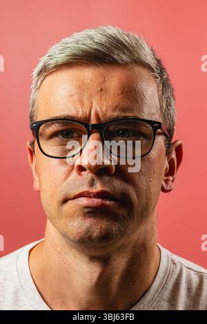 Un uomo serio con capelli grigi corti e occhiali neri fissa direttamente la fotocamera. Mostra un'espressione focalizzata, in contrasto con un pi vibrante Foto Stock