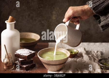 due ciotole di tè matcha con brownie con cacao in polvere sul tavolo, una donna che versa latte dal vaso in ceramica Foto Stock