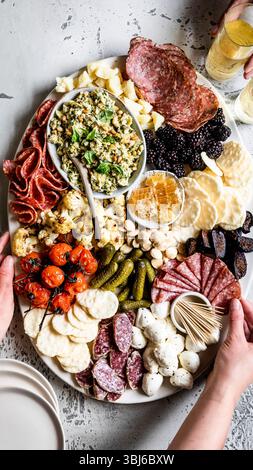 Un tagliere di salumi assortito con carni, formaggi, cracker, frutta, noci, e sottaceti, con una persona che regge un bicchiere di champagne. Foto Stock