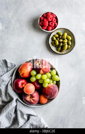 Un assortimento di frutta fresca, tra cui pesche, uva, albicocche, lamponi e olive disposte in ciotole su una superficie ruvida. Foto Stock