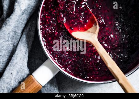 Una pentola ripiena di ricca marmellata di bacche scure e un cucchiaio di legno su sfondo grigio. Foto Stock