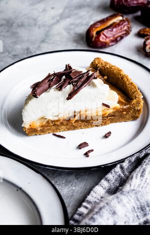 Una fetta di torta condita con panna montata e scaglie di cioccolato su un piatto bianco, con datteri sullo sfondo. Foto Stock