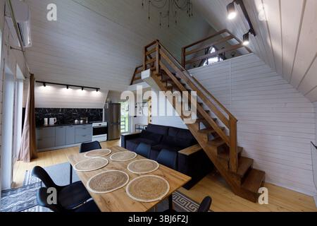 Questo splendido interno presenta una cucina elegante con ripiani scuri, un'accogliente zona pranzo con texture naturali e un'elegante scala in legno Leadi Foto Stock