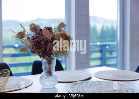 Questa immagine mostra un elegante tavolo da pranzo adornato con una bella disposizione di fiori secchi in un vaso di vetro, completato da uno scenario naturale visibile Foto Stock