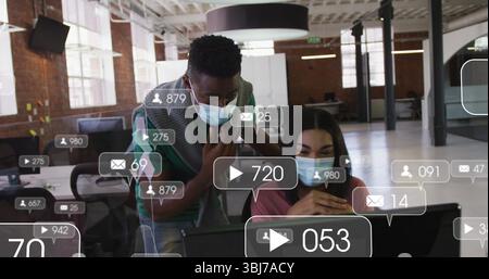 Collaborazione con colleghi mascherati che analizzano le statistiche dei social media su un notebook in ufficio condiviso, icone di dati Foto Stock