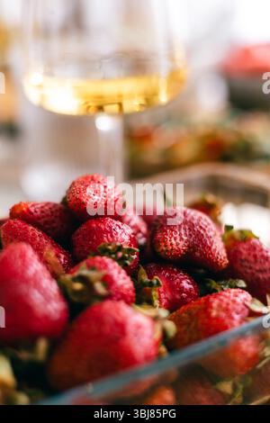 Fragole fresche mature in una ciotola di vetro con bicchiere di vino in fondo morbido, frutta estiva Foto Stock