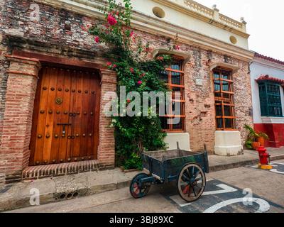 Una strada pittoresca a Cartagena, Colombia, caratterizzata da una storica porta in legno, fiori vivaci e un antico carro contro un muro rustico. Foto Stock