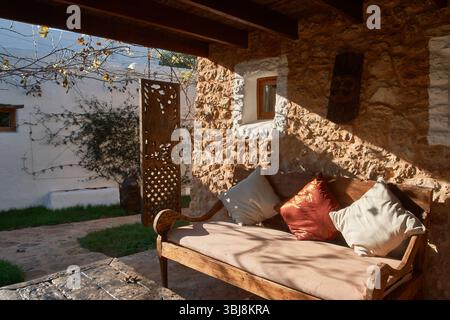 Accogliente panca in legno con cuscini invita al relax su un portico di una casa rustica in pietra, bagnata dalla calda luce del sole Foto Stock