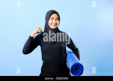 Una donna che indossa un hijab nero regge un tappetino blu per lo yoga. Lei sorride ed è felice Foto Stock