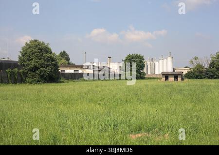 Baracca in cemento abbandonata su un campo con un complesso industriale sullo sfondo in una giornata di sole nella campagna italiana Foto Stock