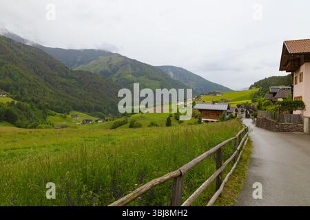 Vista panoramica della Valle di Funes nelle Dolomiti, Italia, con una strada di campagna, prati verdi e case alpine in una giornata nuvolosa. Foto Stock