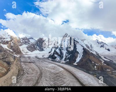Vista aerea di un vasto ghiacciaio che si snoda attraverso una serie di maestose montagne innevate sotto un cielo blu nuvoloso Foto Stock