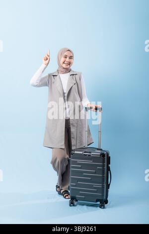 Una donna che indossa un gilet grigio e una camicia bianca regge una valigia nera. Sta indicando qualcosa e sorridendo Foto Stock
