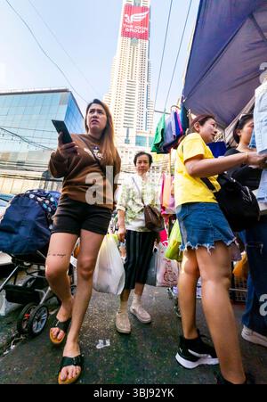 Thailandia - 7 aprile 2024 - molte persone curiosano e comprano merci in un mercato all'aperto pomeridiano, con il Baiyoke Sky Hotel che torreggia alla luce del sole oltre. Foto Stock