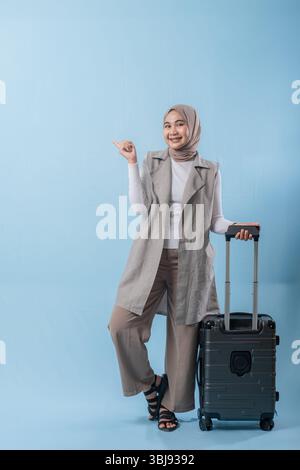 Una donna che indossa una camicia bianca e un gilet grigio tiene in mano una valigia e indica qualcosa di importante Foto Stock