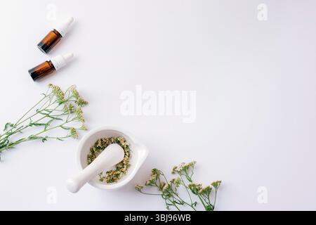 Mortaio e pestello con erbe essiccate e bottiglie di olio essenziale su sfondo bianco con fiori di freccia. Vista dall'alto, disposizione piatta, spazio di copia. Foto Stock