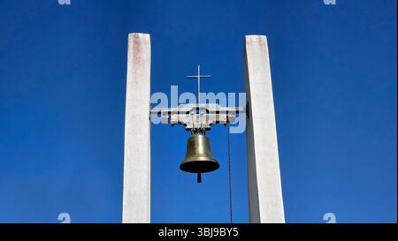 Il campanile bianco minimalista con campana in ottone e croce in metallo si erge alto contro un cielo blu profondo, simboleggiando la fede e la semplicità in stile contemporaneo Foto Stock
