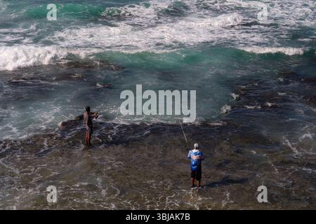 Guardando dall'alto verso il basso due uomini che pescano dalle rocce con i piedi in acqua sulla riva di una spiaggia sulla costa di Durban, in Sudafrica. Foto Stock