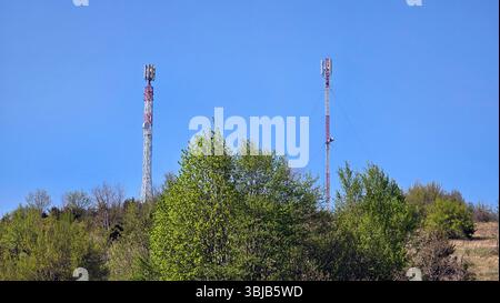 Due alte torri cellulari con antenne e strutture a reticolo rosso-bianco sorgono sopra una collina di alberi primaverili frondosi, adagiati contro un cielo blu senza nuvole Foto Stock