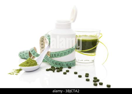 Polvere di erba, pillole di spirulina verde, vetro con agitazione vegetale verde e metro a nastro isolato su sfondo bianco. Salute verde abbraccio Foto Stock