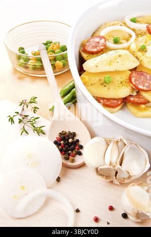 Fette di patate fresche con verdure fresche e salsiccia preparate per la cottura nel recipiente su un tavolo di legno Foto Stock