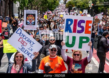 San Francisco, Stati Uniti. 14 giugno 2025. I manifestanti sorridenti reggono cartelli che recitano: "La democrazia muore in silenzio", "RISE UP" e versi dei musical Hamilton e Les Miserables: "Oceans Rise, Empires Fall" e "Do you hear the People sing?" Dietro di loro migliaia di manifestanti riempiono la strada nella diestenza. Crediti: Shelly Rivoli/Alamy Live News Foto Stock