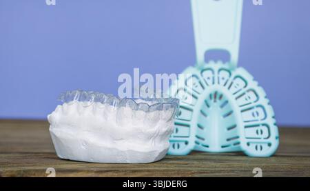 Allineatore ortodontico trasparente posizionato su uno stampo dentale in gesso, con un vassoio di impronta sullo sfondo su una superficie in legno. Foto Stock
